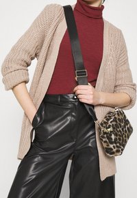 Beige chunky gebreide cardigan over een bordeauxrode coltrui, gecombineerd met hoge zwarte neplederen broeken en een kleine handtas met luipaardprint.