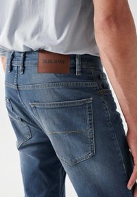 Jeans in denim blu scuro con una texture liscia, caratterizzati da un'etichetta in pelle marrone marchiata e cuciture visibili sulle tasche posteriori.