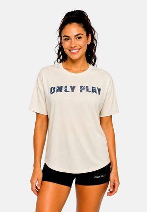Lächelnde Frau mit dunklen Haaren im Pferdeschwanz, trägt ein beiges "ONLY PLAY" T-Shirt und schwarze Shorts mit "ONLY PLAY" Text am linken Bein.