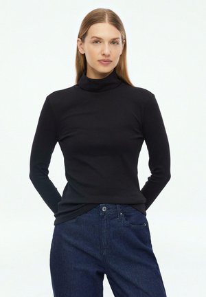 Woman with straight light brown hair wearing a black turtleneck sweater and dark blue jeans, standing with hands behind her back.