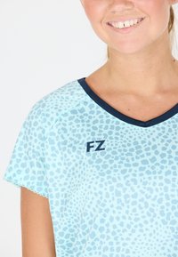 Kvinna som ler, iklädd en ljusblå skjorta med marinblå detaljer och ett abstrakt prickigt mönster, med "FZ"-logotypen på bröstet.