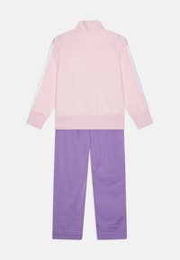 To-delt tracksuit: Lys pink zip-up jakke med høj krave, hvide striber i siderne; lilla bukser med afslappet pasform og glat tekstur.