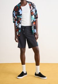 Chemise à manches courtes, à motif coloré, avec un short noir. Chaussures noires à enfiler et chaussettes blanches. Tissu lisse, coupe décontractée, motifs géométriques vibrants.