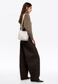 Bolso tipo cubo de piel blanca con forma estructurada y asa corta, adornado con una marca grabada. Combinado con un suéter marrón de felpa y pantalones oscuros.