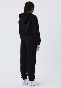 Zwarte hoodie en bijpassende joggers met een cuff aan de enkels, gemaakt van zachte stof. Gdracht met witte sneakers. Achteraanzicht tegen een lichte achtergrond.
