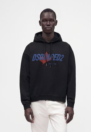Homme portant un sweat à capuche noir DSQUARED2 avec un logo feuille d'érable rouge et un pantalon blanc, debout devant un fond clair uni.
