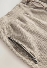 Beżowe spodnie z tkaniny plecionej, z teksturowanym wzorem, posiadające boczną kieszeń na zamek, elastyczny pas ze sznurkiem oraz gładką podszewkę.