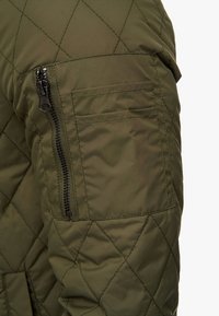 Ärmels eines olivgrünen gesteppte Jacke mit einer Reißverschlusstasche und strukturiertem Stoff. Die Jacke zeigt ein durchgehendes Diamantmuster.