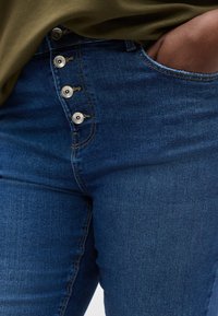 Άτομο που φοράει ψηλόμεσα μπλε τζιν με τέσσερα ορατά κουμπιά, με το χέρι στο εμπρόσθιο τσέπη και ένα χιτώνα σε ελιά πράσινο χρώμα.