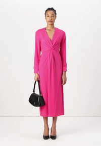 Pink, long-sleeved dress with a deep V-neck and twisted front detail. Worn with black high heels and a black handbag. Smooth fabric.