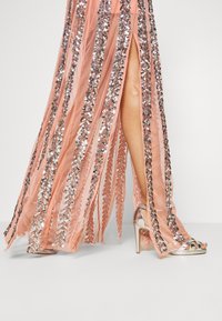 Persikofärgad aftonklänning med vertikala streck av silverpaljetter och ett högskuret slits, kombinerad med metalliska klacksandaler. Slät tygkvalitet.