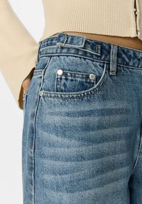 Jeans in denim blu con una texture sbiadita, dotati di un design a cinque tasche e rivetti metallici. La vita include un passante per la cintura.
