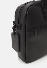 Schwarze synthetische Tasche mit strukturiertem Design, glatter Textur, doppeltem Reißverschluss und einem abnehmbaren Etikett mit Markennamen auf der Seite.