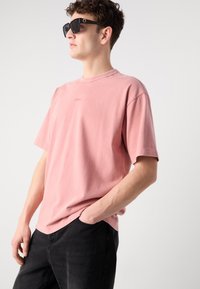 Lyserød bomulds t-shirt med korte ærmer og rund halsudskæring. Har et diskret logo på brystet og er parret med sorte jeans.