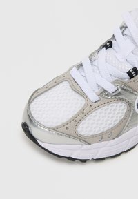 Sneaker z białą siateczką na górze, metalicznie srebrnymi akcentami, fakturowanymi szarymi overlayami i białymi sznurowadłami. Czarna gumowa podeszwa z bieżnikiem.