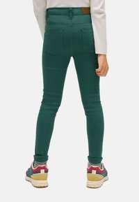 Jeans ajustados verdes con una textura suave, que cuentan con dos bolsillos traseros y una etiqueta de cuero marrón en la cintura, combinados con zapatillas multicolores.