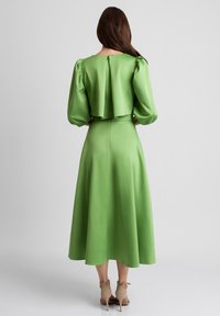 Zielona satynowa sukienka z bufiastymi rękawami, plisowanym tyłem i zwiewną spódnicą. Wyróżnia się gładką teksturą i długością do kolan.