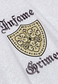 Tissu gris avec un motif de bouclier en or et noir, présentant des motifs floraux et des bordures complexes. Le mot "Infame" est brodé en police sombre.
