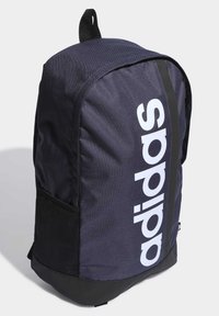 Mochila de tela azul marino con gran logotipo blanco de "adidas", forma curva, fondo negro y bolsillo lateral de malla. Cuenta con un asa en la parte superior para llevar.