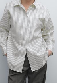 Persona che indossa una camicia bianca a maniche lunghe con strisce verticali e mani nelle tasche, abbinata a pantaloni grigi.