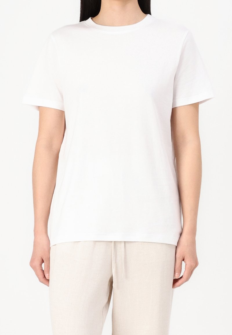 Personne portant un t-shirt blanc uni à manches courtes et un pantalon beige clair, debout devant un fond blanc.