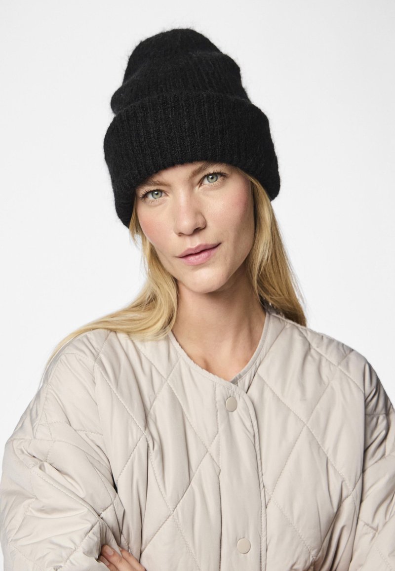 Fekete kötött sapka hajtott peremmel, puha gyapjúból, világos bézs színű, rombuszmintás steppelt dzsekihez párosítva, patentgombokkal.