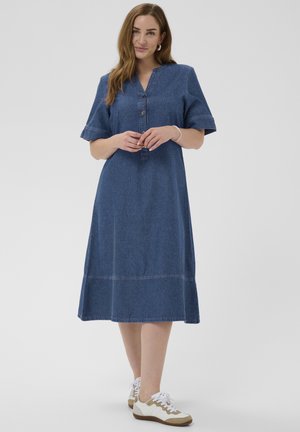 Kvinde stående iført en blå denim midi-kjole med korte ærmer og hvide sneakers mod en ens grå baggrund.