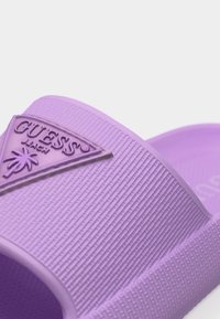 Κοντινό πλάνο του λουριού σανδάλου τύπου slide σε λαβανδί χρώμα με υφή στην επιφάνεια και ανάγλυλο λογότυπο Guess Beach που απεικονίζει έναν φοίνικα.