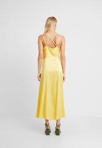 Robe maxi en satin jaune avec un dos nu et des bretelles croisées. Le tissu a une texture lisse, un design fluide et un ourlet jusqu'aux genoux.