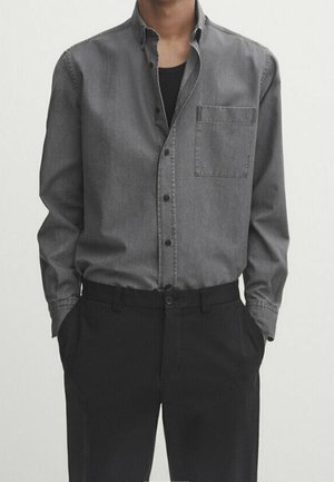 Personne portant une chemise grise boutonnée sur un t-shirt noir, avec un pantalon noir, debout les mains dans les poches devant un fond uni.