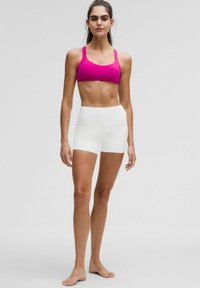 Sostén deportivo de color rosa brillante con tirantes anchos, combinado con pantalones cortos blancos de cintura alta. Textura suave, diseño ajustado y detalles mínimos.