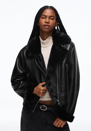 Femme aux longues tresses portant une veste en cuir noire doublée de fausse fourrure, par-dessus un pull à col roulé beige clair et un pantalon noir.