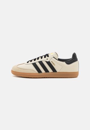 Beiger Adidas Samba Sneaker mit schwarzen Streifen, schwarzen Schnürsenkeln, schwarzer Ferse und gummifarbener Sohle auf weißem einfarbigem Hintergrund.