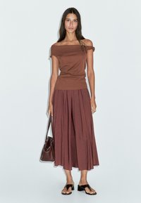 Top marrone scollato abbinato a una lunga gonna plissettata color marrone bordeaux. La modella tiene una borsa marrone e indossa sandali piatti neri. Sfondo semplice.