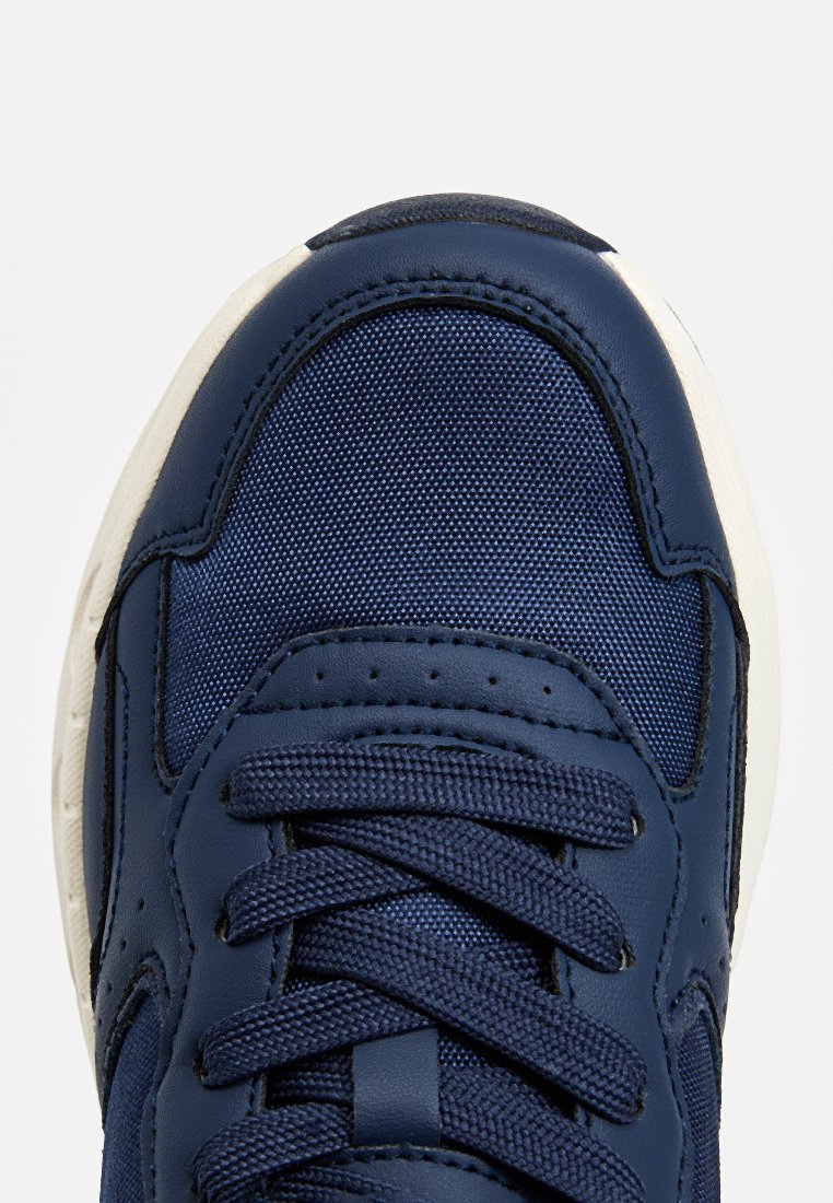 Zapatilla azul marino con parte superior de tela texturizada, acentos de cuero, cordones planos y suela de goma blanca. Presenta un diseño de punta redonda.