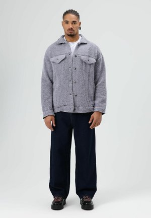 Grijze pluizige jas met twee borstzakken, knoopsluiting en een kraag, gecombineerd met donkerdere wijde jeans en zwarte veterschoenen.