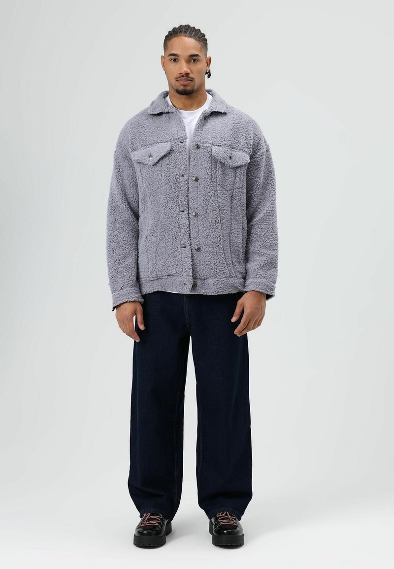 Grijze pluizige jas met twee borstzakken, knoopsluiting en een kraag, gecombineerd met donkerdere wijde jeans en zwarte veterschoenen.