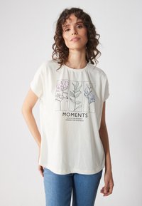 Kreemja värvi lühikeste varrukatega T-särk, millel on minimalistlik lilleline graafiline kujundus ja sõnad "MOMENTS" musta värviga.