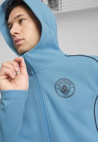 Hættejakke med lynlås i lyseblåt stof med sorte detaljer. Har Manchester City-logoet på brystet. Glat tekstur, atletisk pasform.