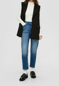 Černá vesta s klopami, bílý žebrovaný svetr s dlouhým rukávem, modré roztrhané džíny a černé mokasíny. Viditelné ohrnuté lemy a kontrast textur.