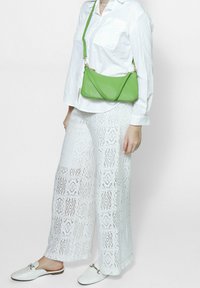 Pantaloni bianchi in pizzo a gamba larga con ritagli decorativi, una camicia bianca aderente con bottoni e una borsa a tracolla in pelle verde. Le scarpe bianche slip-on completano il look.