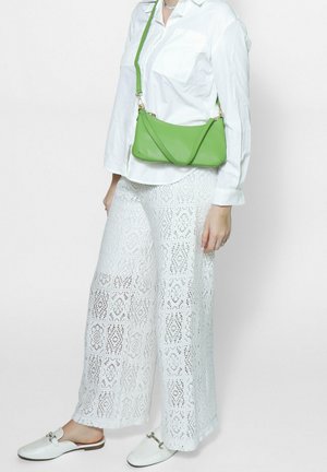 Weiße Spitzen-Hosen mit weitem Bein und gemusterten Ausschnitten, ein tailliertes weißes Hemd mit Knöpfen und eine grüne Leder-Crossbody-Tasche. Weiße Slipper runden das Outfit ab.
