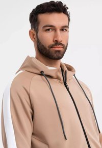 Felpa con zip di colore beige, con cordini neri, accenti bianchi sui lati e una texture morbida. Presenta un proteggi mento e un design aderente.