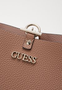 Sac à main en cuir texturé marron avec un logo "GUESS" doré et un accent matériel circulaire argenté en haut.