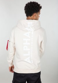 Haut à capuche beige avec un grand logo blanc "ALPHA INDUSTRIES" au dos, dispose d'une capuche, de manches à poignets côtelés et d'un détail de poche rouge sur la manche.
