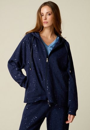 Vrouw die een donkerblauwe pailletten hoodie en bijpassende broek draagt over een lichtblauw overhemd, staand met één hand in haar zak tegen een effen achtergrond.