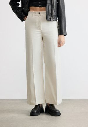 Woman wearing high-waist wide-leg cream trousers, black cropped top, black leather jacket, and chunky black loafers standing on gray floor.