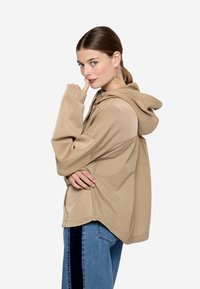 Sudadera beige con capucha, de manga larga, ajuste relajado, cortes laterales y puños acanalados. Se lleva con vaqueros azules con paneles laterales negros.