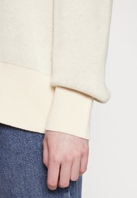 Roomkleurige gebreide trui met geribbelde manchetten en een getextureerd oppervlak. Hand in blauwe denim, die het kleurcontrast van de stof benadrukt.