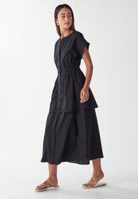 Abito nero a bottoni con maniche corte, vita elastica, design a strati e lunghezza midi. Indossato con sandali piatti con cinturino.